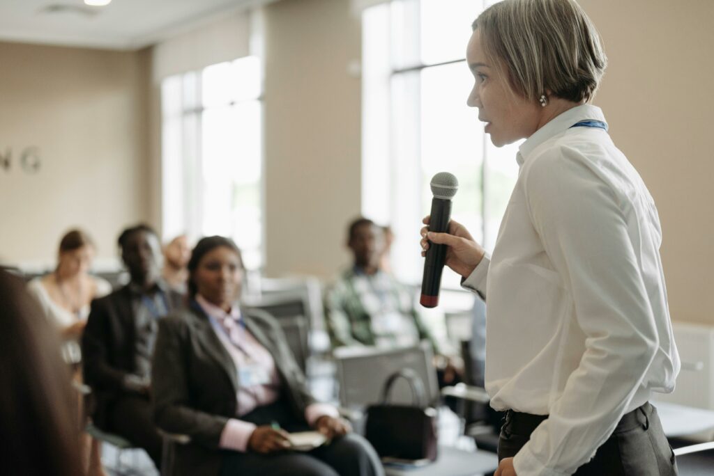 pexels-photo-8761534-8761534 Side view of a businesswoman holding a microphone while addressing an audience at a conference.